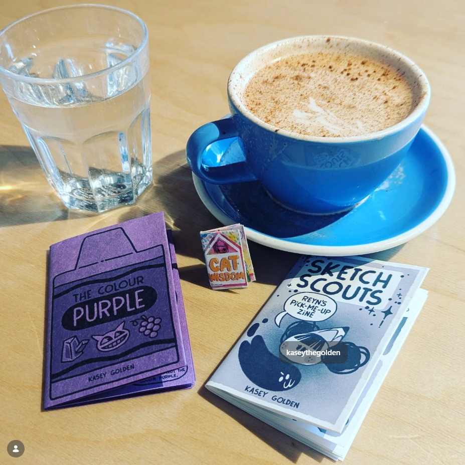 wooden table with a glass of water (upper left), chai latte in a blue mug and saucer (upper right), a purple zine labeled The color Purple by Kasey Golden (bottom left), a especially small zine  in bright pinks and oranges and blues named Cat Wisdom (center), and a blue grey zine named Sketch Scouts: Reyn's Pick-me-up Zine by Kasey Golden (bottom right) 